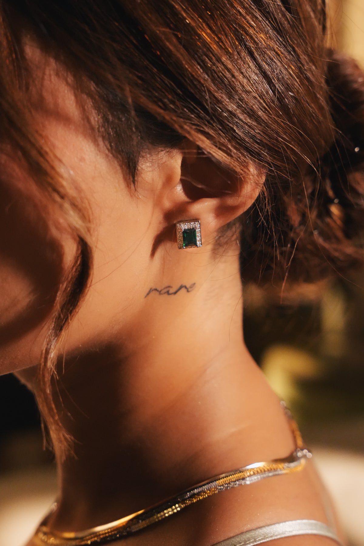 A close-up lifestyle shot of a woman's ear and neck, showcasing a small square green gemstone stud earring with a silver-toned setting. 